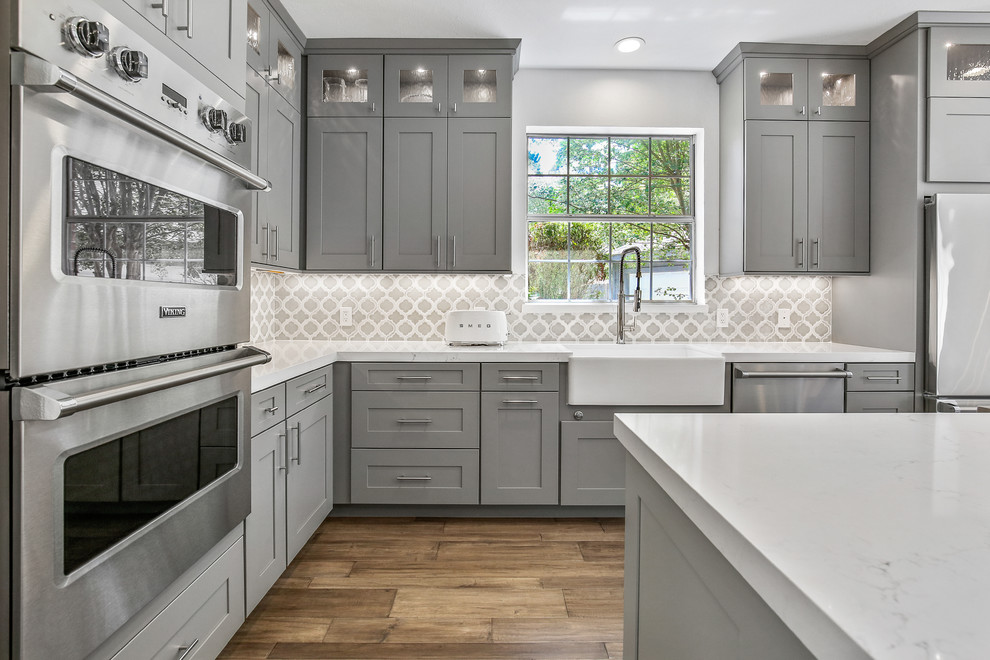 Example of a mid-sized transitional l-shaped light wood floor and brown floor eat-in kitchen design in Houston with a farmhouse sink, shaker cabinets, gray cabinets, quartzite countertops, blue backsplash, porcelain backsplash, stainless steel appliances, an island and white countertops