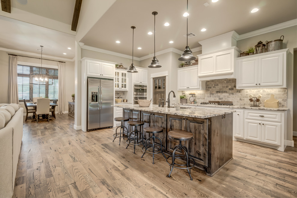 Mid-sized transitional l-shaped medium tone wood floor open concept kitchen photo in Oklahoma City with an undermount sink, raised-panel cabinets, white cabinets, granite countertops, gray backsplash, brick backsplash, stainless steel appliances and an island