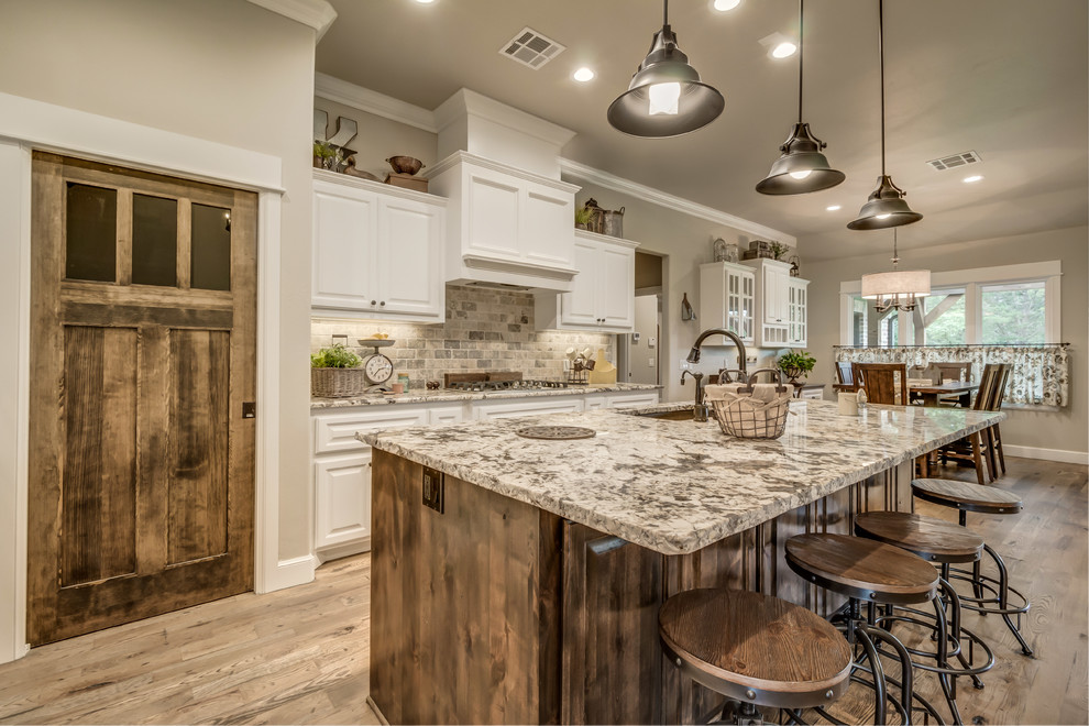 Open concept kitchen - mid-sized farmhouse l-shaped medium tone wood floor open concept kitchen idea in Oklahoma City with an undermount sink, raised-panel cabinets, white cabinets, granite countertops, gray backsplash, brick backsplash, stainless steel appliances and an island