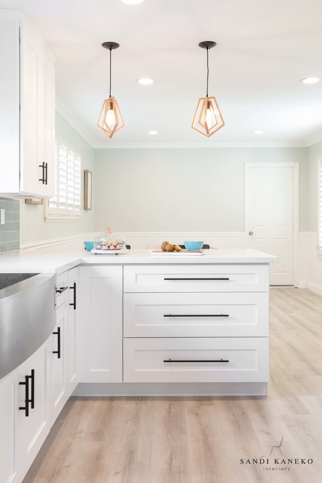 Photo of a small modern galley kitchen/diner in Los Angeles with a belfast sink, shaker cabinets, white cabinets, engineered stone countertops, blue splashback, glass tiled splashback, stainless steel appliances, light hardwood flooring, a breakfast bar, grey floors and white worktops.