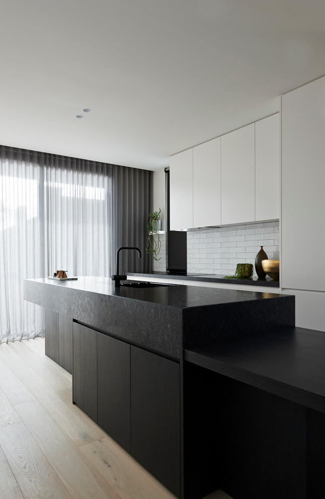 Inspiration for a contemporary single-wall kitchen/diner in Melbourne with a built-in sink, flat-panel cabinets, black cabinets, granite worktops, white splashback, metro tiled splashback, light hardwood flooring and an island.