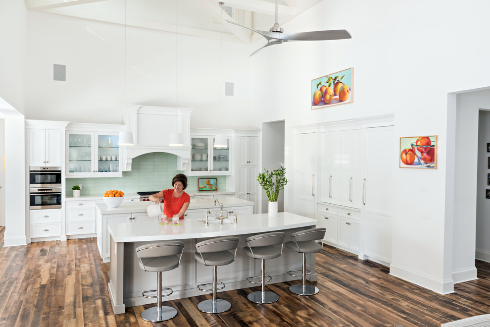 This is an example of a nautical kitchen in Miami with glass-front cabinets, white cabinets, green splashback, glass tiled splashback, stainless steel appliances, dark hardwood flooring, multiple islands and brown floors.