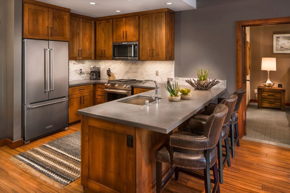 Example of a mid-sized mountain style u-shaped medium tone wood floor and brown floor open concept kitchen design in Other with an undermount sink, shaker cabinets, medium tone wood cabinets, solid surface countertops, white backsplash, marble backsplash, stainless steel appliances and a peninsula