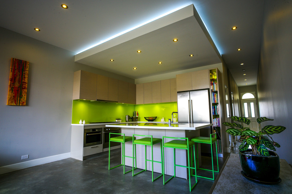 Example of a small trendy l-shaped concrete floor eat-in kitchen design in Sydney with flat-panel cabinets, light wood cabinets, green backsplash, stainless steel appliances, an undermount sink, solid surface countertops, glass sheet backsplash and an island