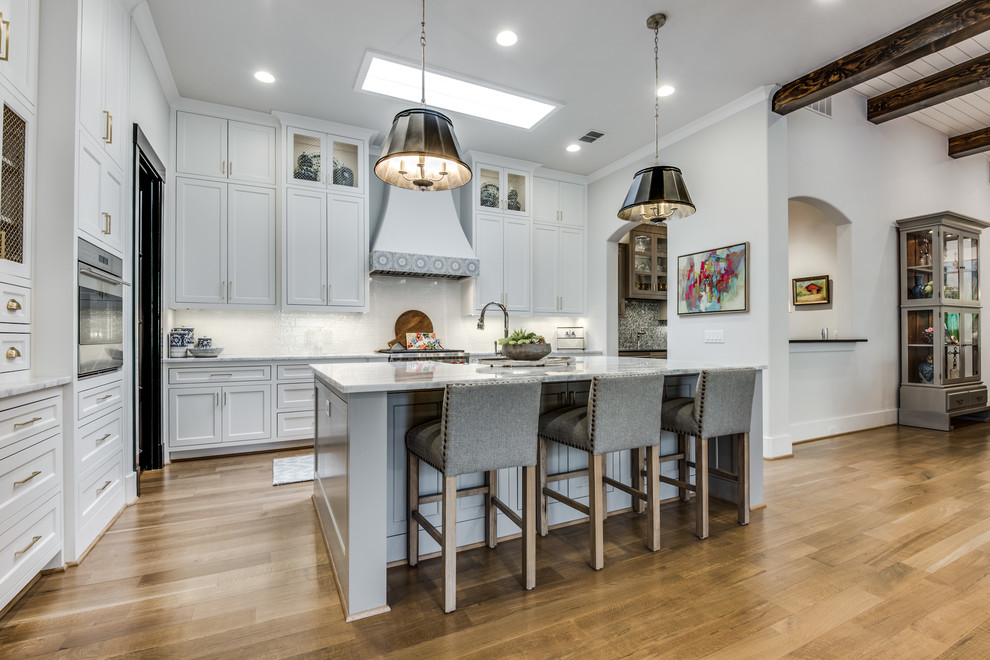 Eat-in kitchen - mid-sized cottage single-wall medium tone wood floor and brown floor eat-in kitchen idea in Dallas with a farmhouse sink, recessed-panel cabinets, white cabinets, marble countertops, white backsplash, marble backsplash, stainless steel appliances and an island