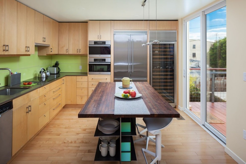 Bild på ett funkis l-kök, med en nedsänkt diskho, släta luckor, skåp i ljust trä, grönt stänkskydd, rostfria vitvaror, ljust trägolv och en köksö