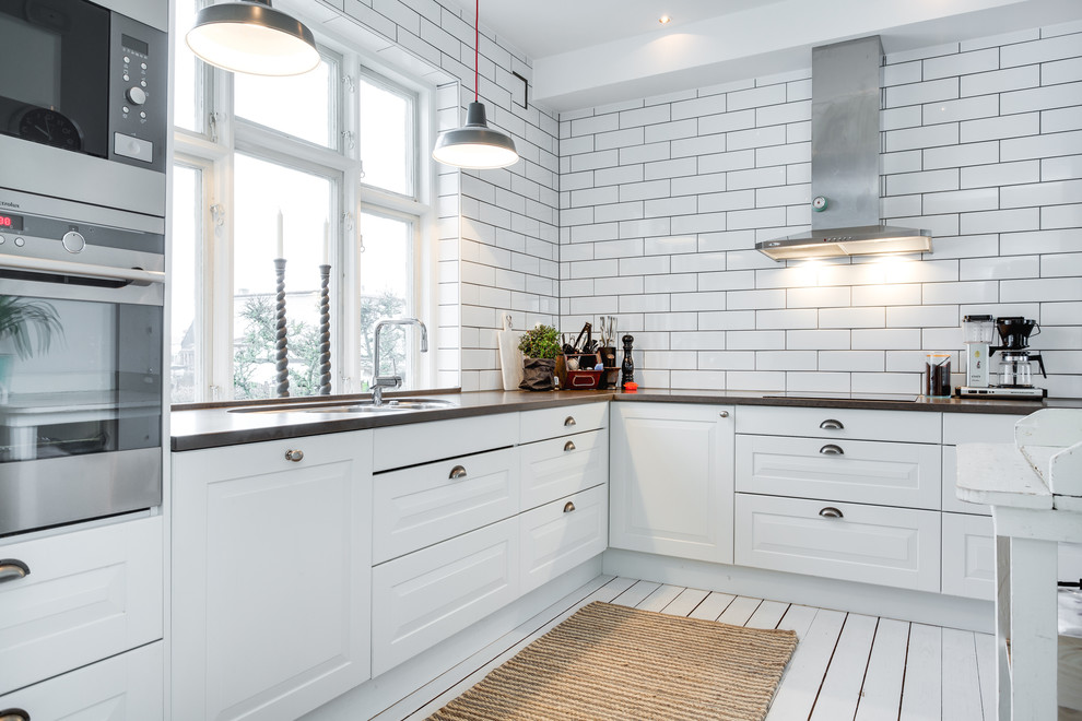 This is an example of a large scandinavian l-shaped enclosed kitchen in Malmo with a double-bowl sink, raised-panel cabinets, white cabinets, wood worktops, stainless steel appliances, painted wood flooring and no island.