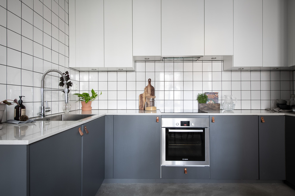 Nordische Küche in U-Form mit Waschbecken, flächenbündigen Schrankfronten, grauen Schränken, Küchenrückwand in Weiß, Küchengeräten aus Edelstahl, Betonboden, grauem Boden und weißer Arbeitsplatte in Göteborg