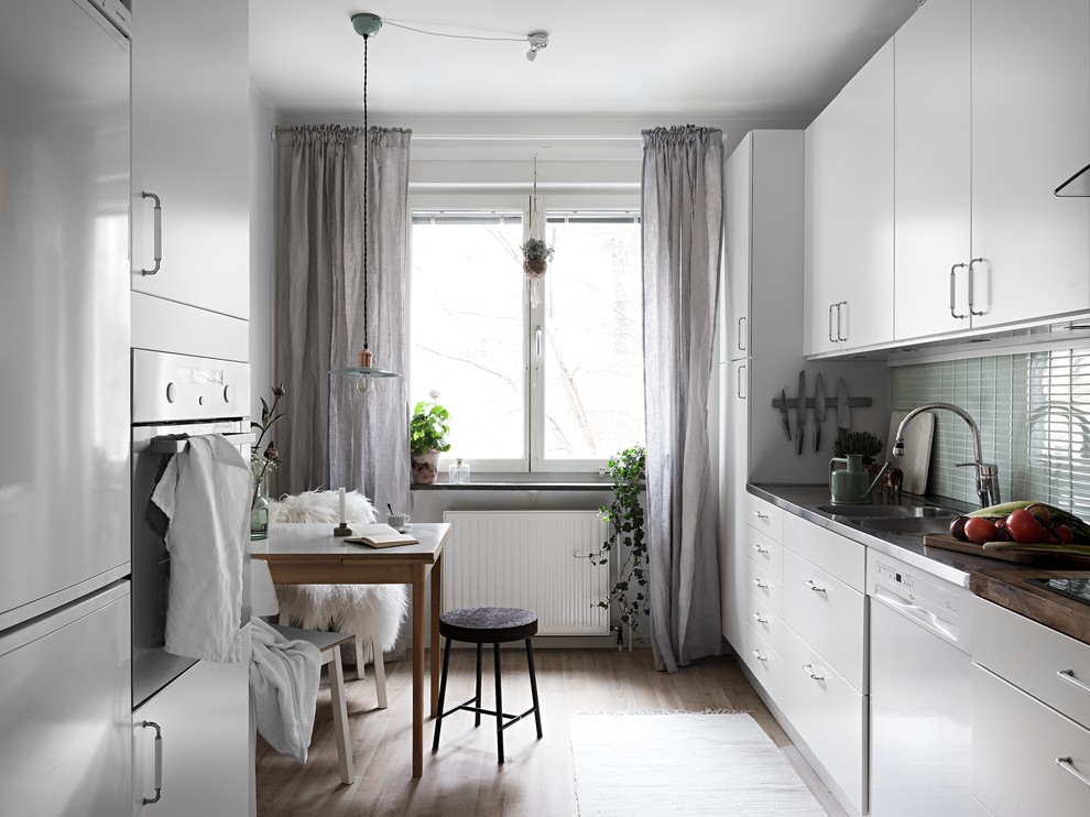 Bild på ett mellanstort skandinaviskt kök, med en dubbel diskho, släta luckor, vita skåp, bänkskiva i rostfritt stål, grönt stänkskydd, stänkskydd i glaskakel, vita vitvaror och ljust trägolv