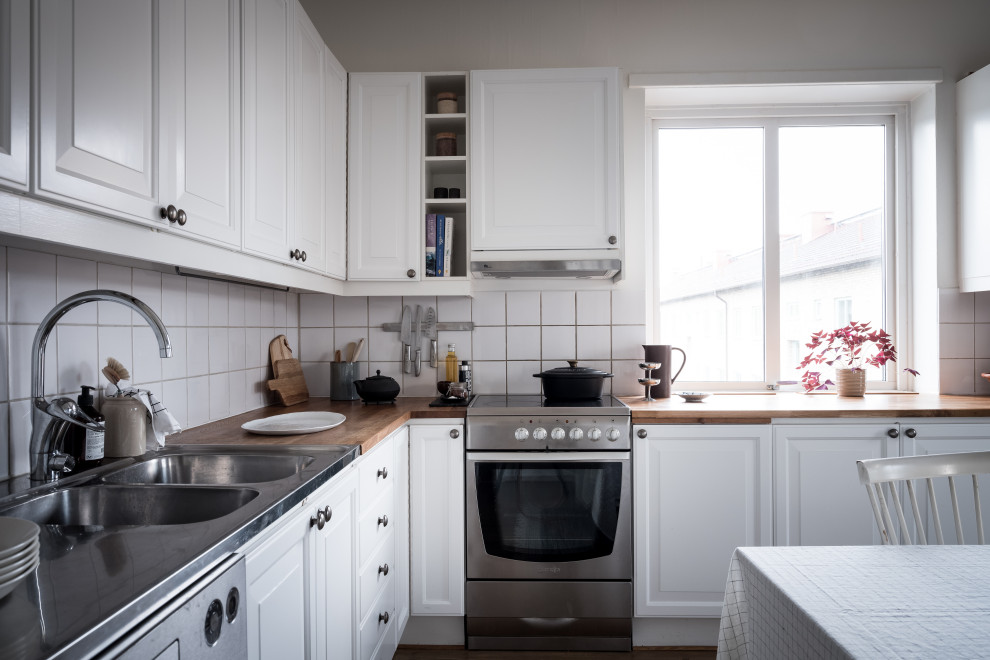 Idéer för ett mellanstort skandinaviskt beige l-kök, med en dubbel diskho, luckor med upphöjd panel, vita skåp, träbänkskiva, vitt stänkskydd, stänkskydd i porslinskakel och rostfria vitvaror