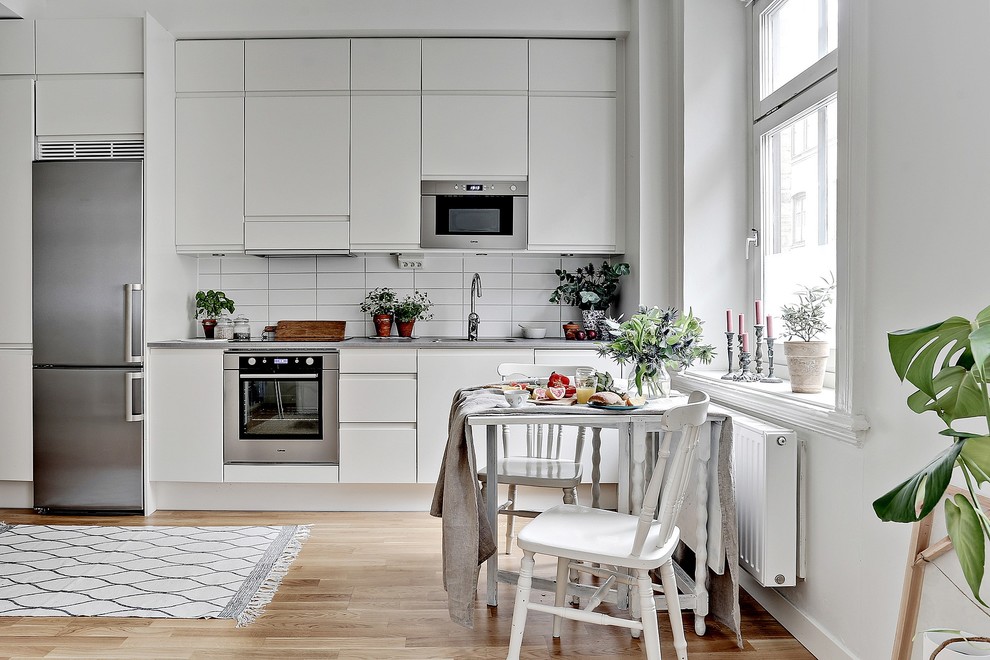 Diseño de cocina comedor lineal escandinava de tamaño medio sin isla con armarios con paneles lisos, puertas de armario blancas, salpicadero blanco, electrodomésticos de acero inoxidable y suelo de madera clara