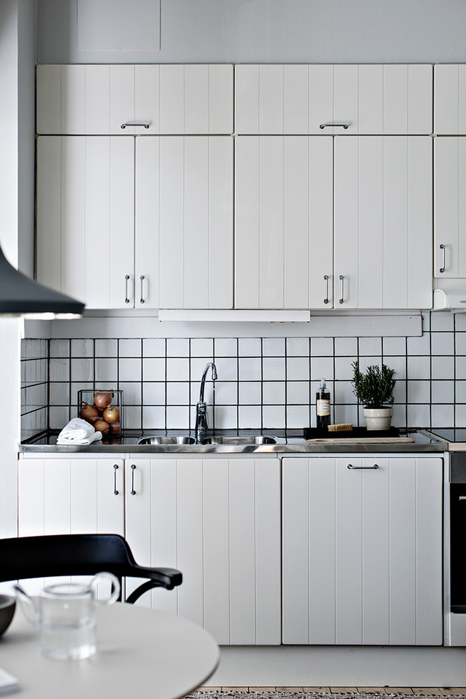 Bild på ett litet minimalistiskt linjärt kök, med en dubbel diskho, luckor med upphöjd panel, vita skåp, bänkskiva i rostfritt stål, vitt stänkskydd och stänkskydd i porslinskakel