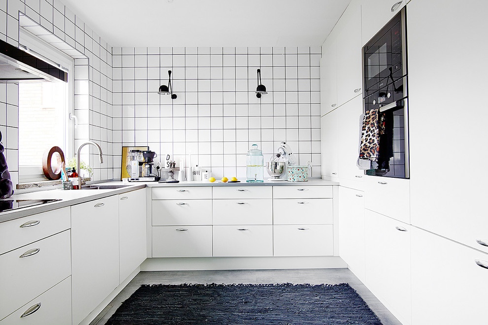 Nordisk inredning av ett mellanstort u-kök, med en undermonterad diskho, släta luckor, vita skåp, vitt stänkskydd, stänkskydd i keramik, svarta vitvaror och betonggolv
