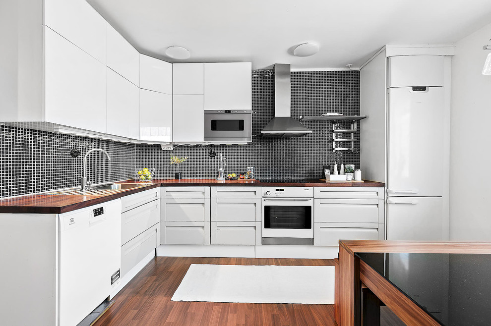 ストックホルムにある北欧スタイルのおしゃれなL型キッチン (フラットパネル扉のキャビネット、白いキャビネット、黒いキッチンパネル、白い調理設備、淡色無垢フローリング) の写真