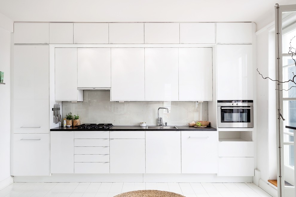 Exempel på ett stort minimalistiskt linjärt kök, med en enkel diskho, släta luckor, vita skåp, grått stänkskydd, glaspanel som stänkskydd, målat trägolv, träbänkskiva och vita vitvaror