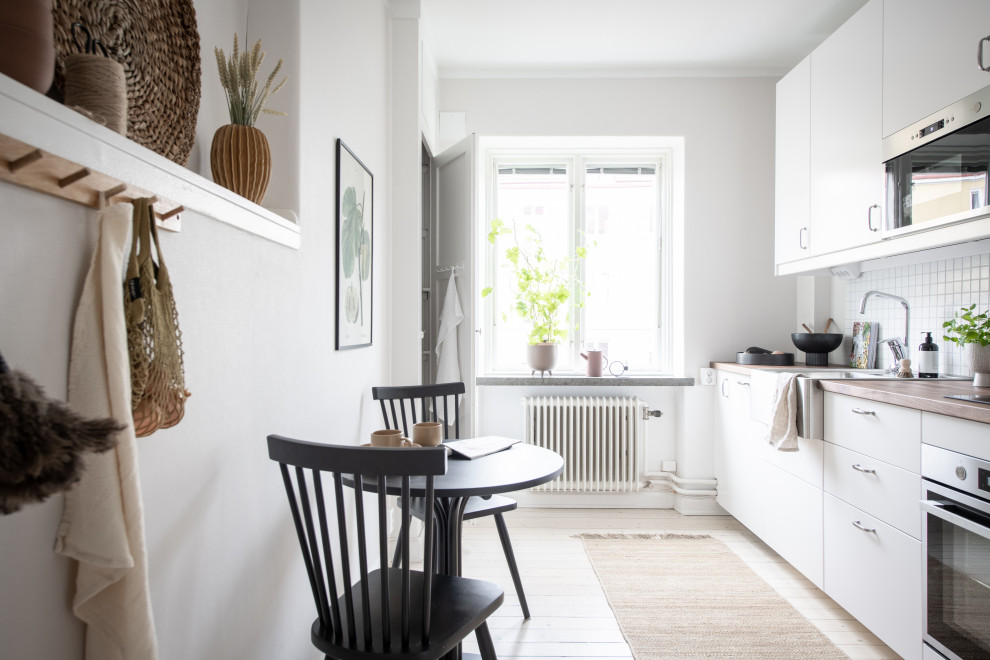 Bild på ett skandinaviskt beige linjärt beige kök och matrum, med en nedsänkt diskho, släta luckor, vita skåp, träbänkskiva, vitt stänkskydd, rostfria vitvaror, ljust trägolv och beiget golv