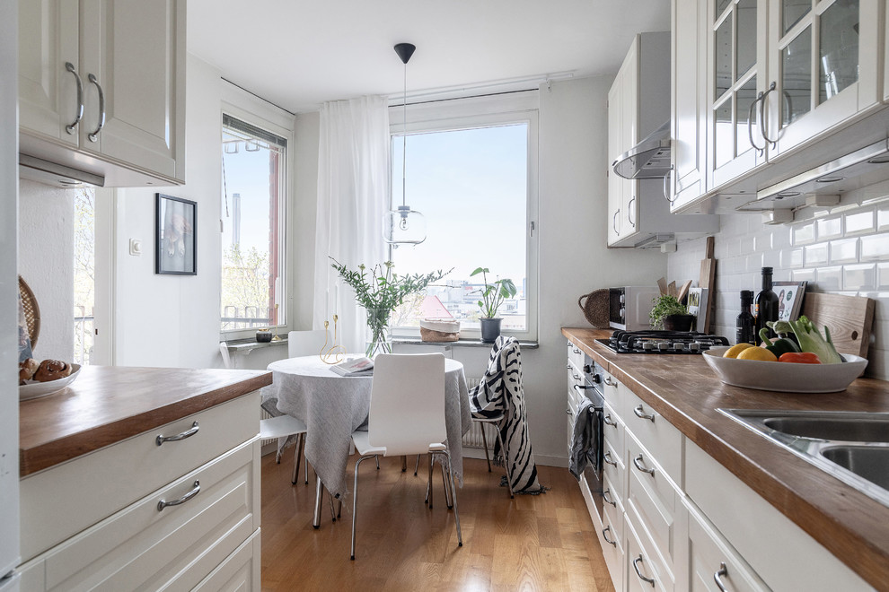 Zweizeilige Skandinavische Wohnküche mit Doppelwaschbecken, weißen Schränken, Arbeitsplatte aus Holz, Küchenrückwand in Weiß, Rückwand aus Metrofliesen und hellem Holzboden in Stockholm