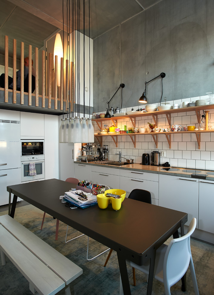 Foto på ett litet industriellt kök och matrum, med släta luckor, vita skåp, vitt stänkskydd, vita vitvaror och betonggolv