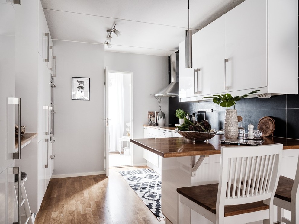 This is an example of a scandi l-shaped kitchen in Stockholm with flat-panel cabinets, white cabinets, wood worktops, black splashback, medium hardwood flooring, a breakfast bar, brown floors and brown worktops.