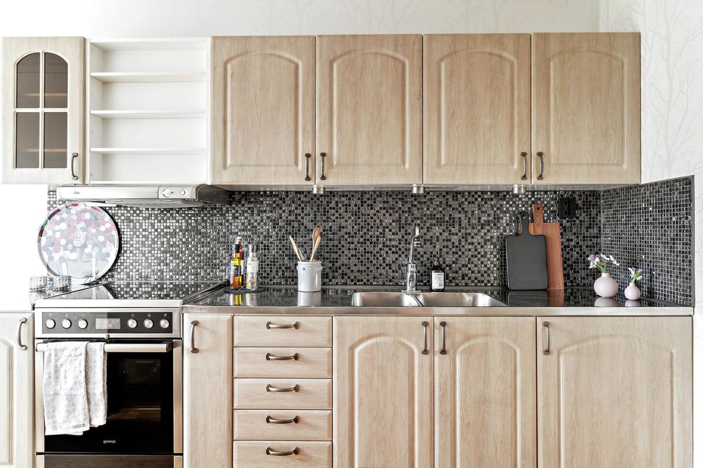 Nordisk inredning av ett mellanstort grå linjärt grått kök, med bänkskiva i rostfritt stål, svart stänkskydd, en integrerad diskho, luckor med upphöjd panel, skåp i ljust trä, stänkskydd i mosaik och rostfria vitvaror