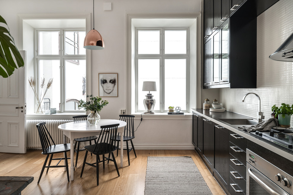 Example of a danish single-wall light wood floor and beige floor eat-in kitchen design in Gothenburg with shaker cabinets, black cabinets, white backsplash, mosaic tile backsplash, stainless steel appliances and gray countertops