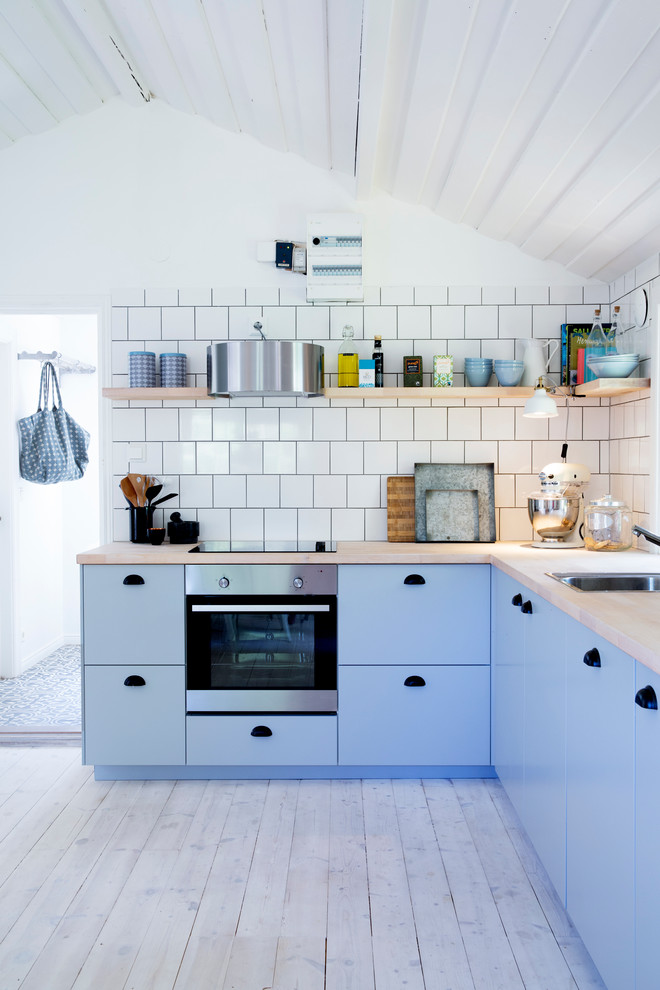 На фото: угловая кухня среднего размера в скандинавском стиле с одинарной мойкой, плоскими фасадами, синими фасадами, деревянной столешницей, техникой из нержавеющей стали, деревянным полом и белым фартуком без острова