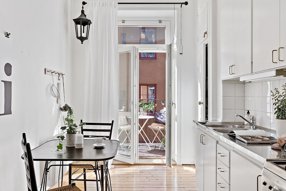 Einzeilige Skandinavische Wohnküche ohne Insel mit flächenbündigen Schrankfronten, weißen Schränken, Edelstahl-Arbeitsplatte, Küchenrückwand in Weiß und hellem Holzboden in Stockholm