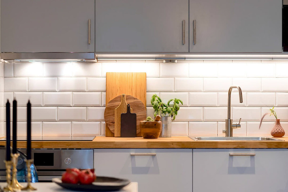 Bild på ett litet funkis linjärt kök med öppen planlösning, med en enkel diskho, släta luckor, grå skåp, träbänkskiva, vitt stänkskydd, stänkskydd i keramik, rostfria vitvaror och mellanmörkt trägolv