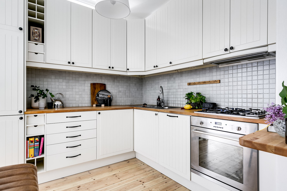 Inspiration for a small scandinavian l-shaped open plan kitchen in Stockholm with white cabinets, wood worktops, grey splashback, light hardwood flooring, no island, a built-in sink, stainless steel appliances, beige floors, beige worktops and beaded cabinets.