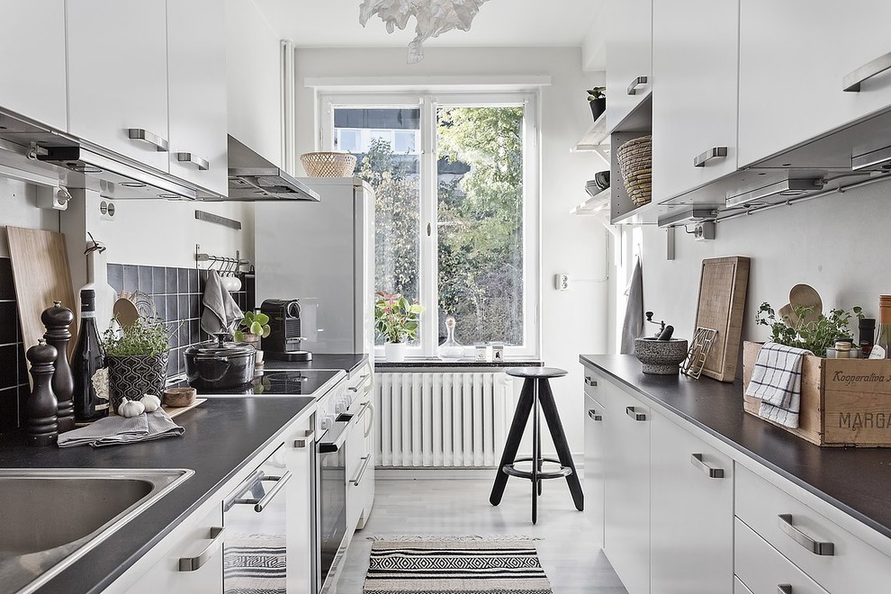 Стильный дизайн: маленькая, узкая отдельная, параллельная кухня в скандинавском стиле с плоскими фасадами, гранитной столешницей, черным фартуком, белой техникой, серым полом и черно-белыми фасадами без острова для на участке и в саду - последний тренд