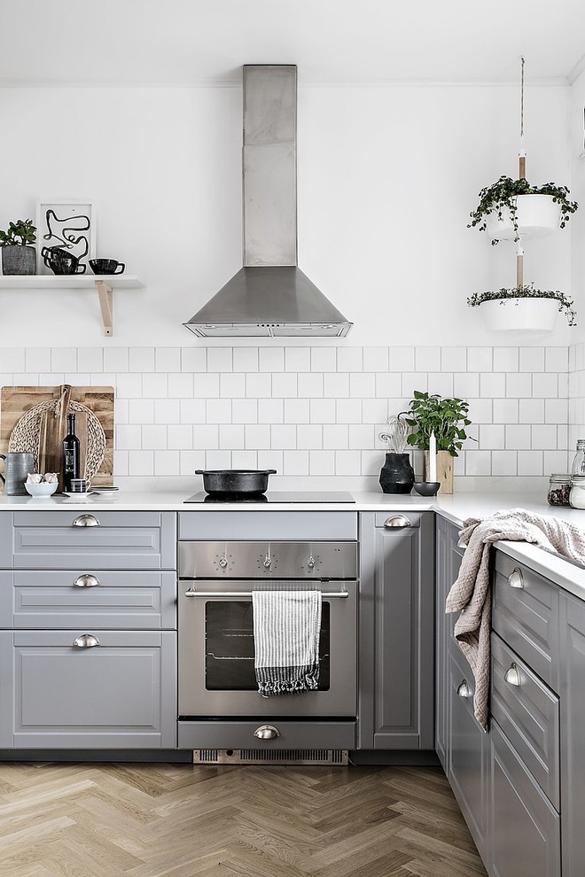 Bild på ett skandinaviskt l-kök, med luckor med upphöjd panel, grå skåp, vitt stänkskydd, stänkskydd i tunnelbanekakel, rostfria vitvaror, ljust trägolv och brunt golv