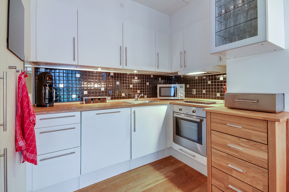 Small contemporary l-shaped enclosed kitchen in Copenhagen with a built-in sink, flat-panel cabinets, white cabinets, wood worktops, black splashback, stainless steel appliances, light hardwood flooring, no island and mosaic tiled splashback.