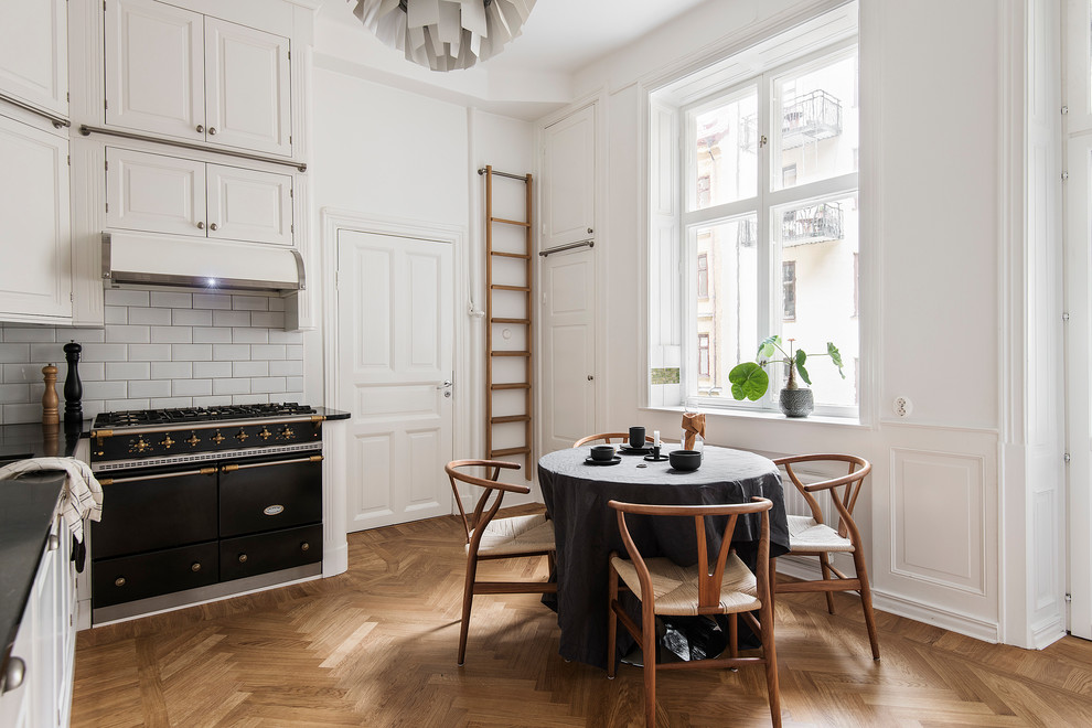 Example of a danish l-shaped medium tone wood floor eat-in kitchen design in Stockholm with raised-panel cabinets, beige cabinets, white backsplash, subway tile backsplash, black appliances and black countertops