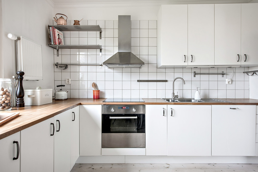 Inredning av ett skandinaviskt l-kök, med en nedsänkt diskho, släta luckor, vita skåp, träbänkskiva, vitt stänkskydd, rostfria vitvaror, ljust trägolv och grått golv