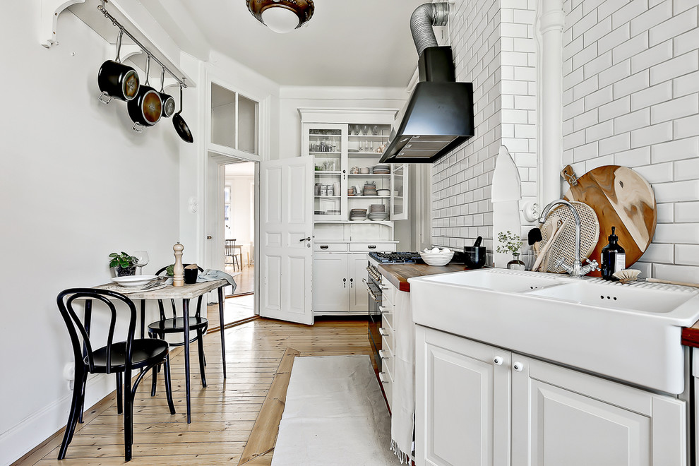 Exempel på ett avskilt, mellanstort nordiskt linjärt kök, med en rustik diskho, luckor med upphöjd panel, vita skåp, träbänkskiva, vitt stänkskydd, stänkskydd i tunnelbanekakel, vita vitvaror och ljust trägolv