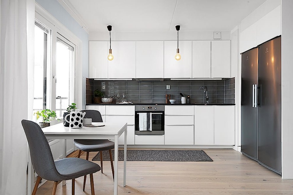 Aménagement d'une cuisine américaine linéaire scandinave avec un placard à porte plane, des portes de placard blanches, une crédence noire, un électroménager en acier inoxydable, parquet clair, un sol beige et plan de travail noir.