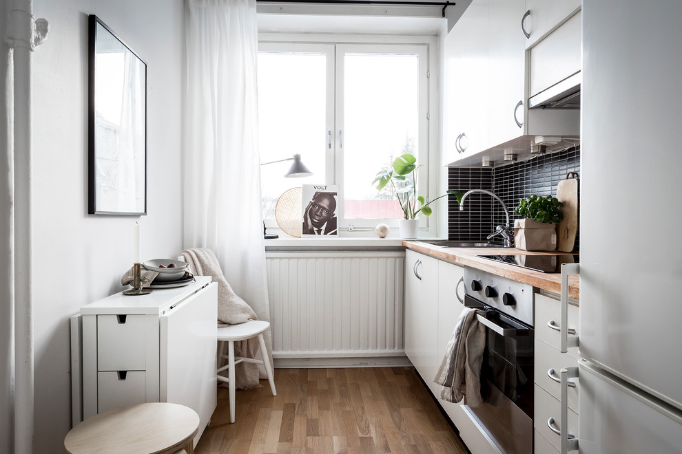 Exempel på ett litet minimalistiskt linjärt kök och matrum, med släta luckor, vita skåp, träbänkskiva, svart stänkskydd, stänkskydd i stickkakel, rostfria vitvaror och mellanmörkt trägolv