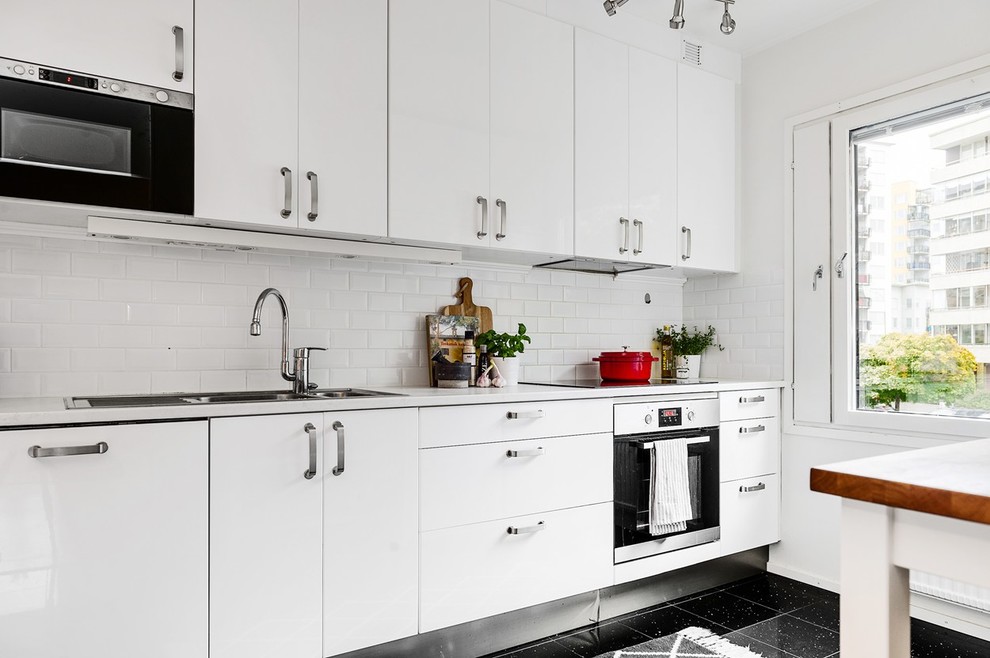 This is an example of a medium sized scandi single-wall open plan kitchen in Stockholm with a double-bowl sink, flat-panel cabinets, white cabinets, white splashback, metro tiled splashback, black appliances and no island.