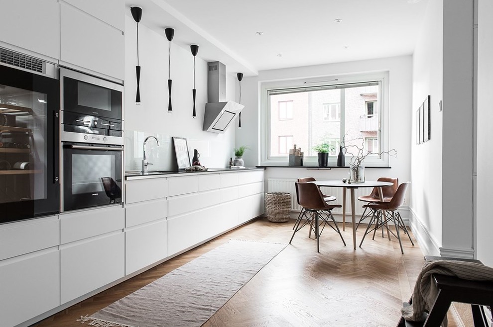 Einzeilige, Mittelgroße Skandinavische Wohnküche ohne Insel mit flächenbündigen Schrankfronten, weißen Schränken, Küchengeräten aus Edelstahl und hellem Holzboden in Göteborg
