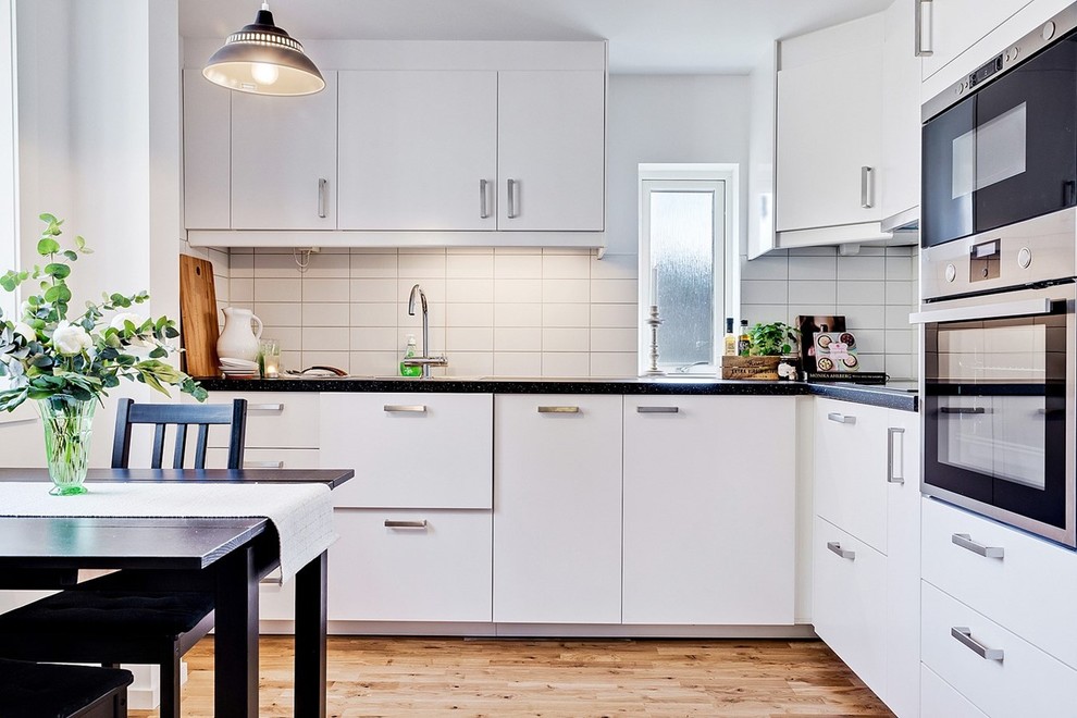 Bild på ett mellanstort nordiskt kök, med släta luckor, vita skåp, vitt stänkskydd, rostfria vitvaror, ljust trägolv och granitbänkskiva