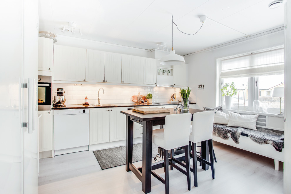 Foto på ett nordiskt linjärt kök och matrum, med en dubbel diskho, beige skåp, vitt stänkskydd, stänkskydd i tunnelbanekakel, vita vitvaror, ljust trägolv, en köksö och vitt golv