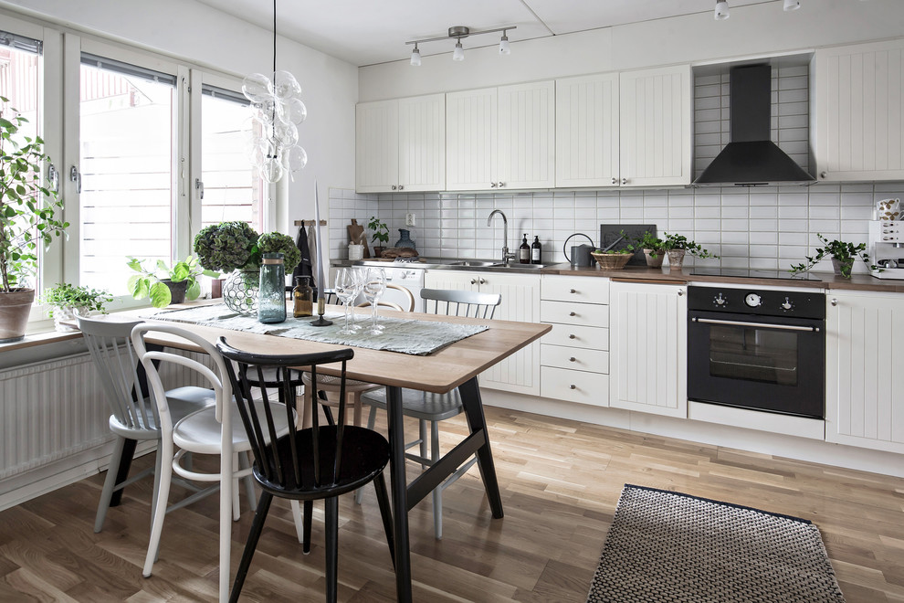 Inspiration for a mid-sized scandinavian single-wall medium tone wood floor and brown floor eat-in kitchen remodel in Gothenburg with a double-bowl sink, white cabinets, wood countertops, white backsplash, black appliances and no island