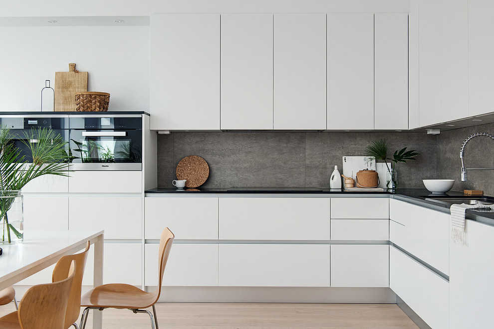 Idée de décoration pour une cuisine américaine nordique en L de taille moyenne avec un placard à porte plane, des portes de placard blanches, un plan de travail en granite, une crédence grise, parquet clair, aucun îlot et un électroménager noir.