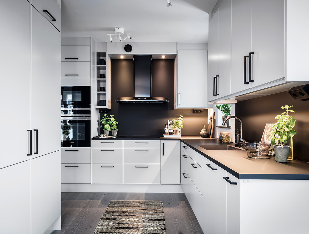 Inredning av ett minimalistiskt stort kök, med en integrerad diskho, släta luckor, vita skåp, svart stänkskydd, svarta vitvaror, grått golv och ljust trägolv