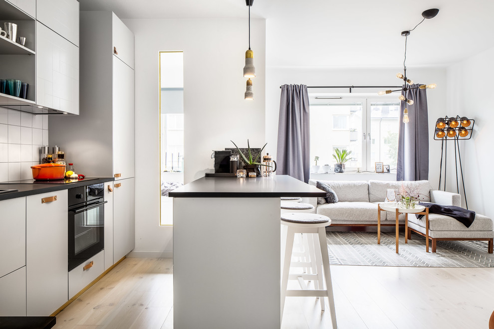 Bild på ett mellanstort skandinaviskt linjärt kök med öppen planlösning, med släta luckor, vita skåp, vitt stänkskydd, svarta vitvaror, ljust trägolv och en halv köksö