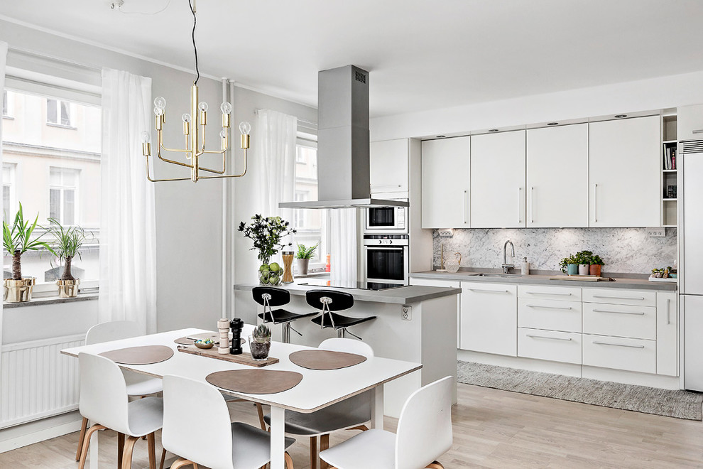 Exempel på ett minimalistiskt kök, med en undermonterad diskho, släta luckor, vita skåp, grått stänkskydd, stänkskydd i sten, rostfria vitvaror, ljust trägolv, en halv köksö och beiget golv