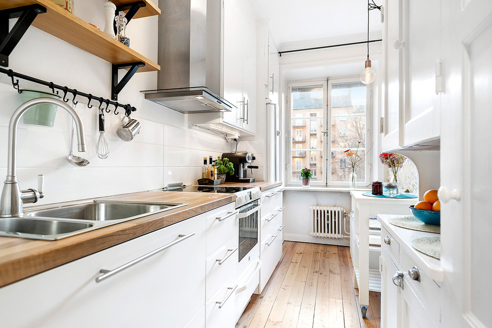 Photo of a medium sized scandinavian single-wall kitchen in Stockholm with a double-bowl sink, flat-panel cabinets, white cabinets, wood worktops, white splashback, stainless steel appliances, light hardwood flooring and no island.