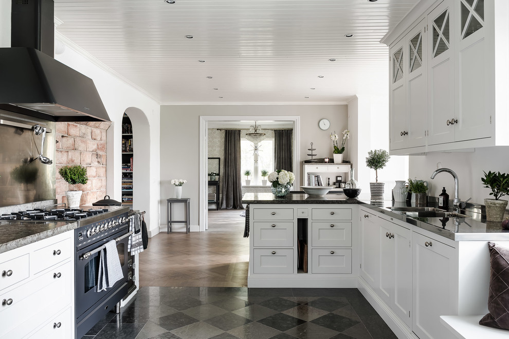 Lantlig inredning av ett stort kök, med en dubbel diskho, luckor med infälld panel, vita skåp, svarta vitvaror, en halv köksö, marmorbänkskiva och stänkskydd med metallisk yta