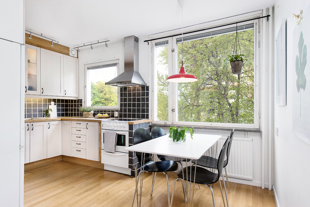Mittelgroße Nordische Wohnküche ohne Insel in L-Form mit weißen Schränken, Arbeitsplatte aus Holz, Küchenrückwand in Schwarz, weißen Elektrogeräten, hellem Holzboden und Rückwand aus Porzellanfliesen in Stockholm
