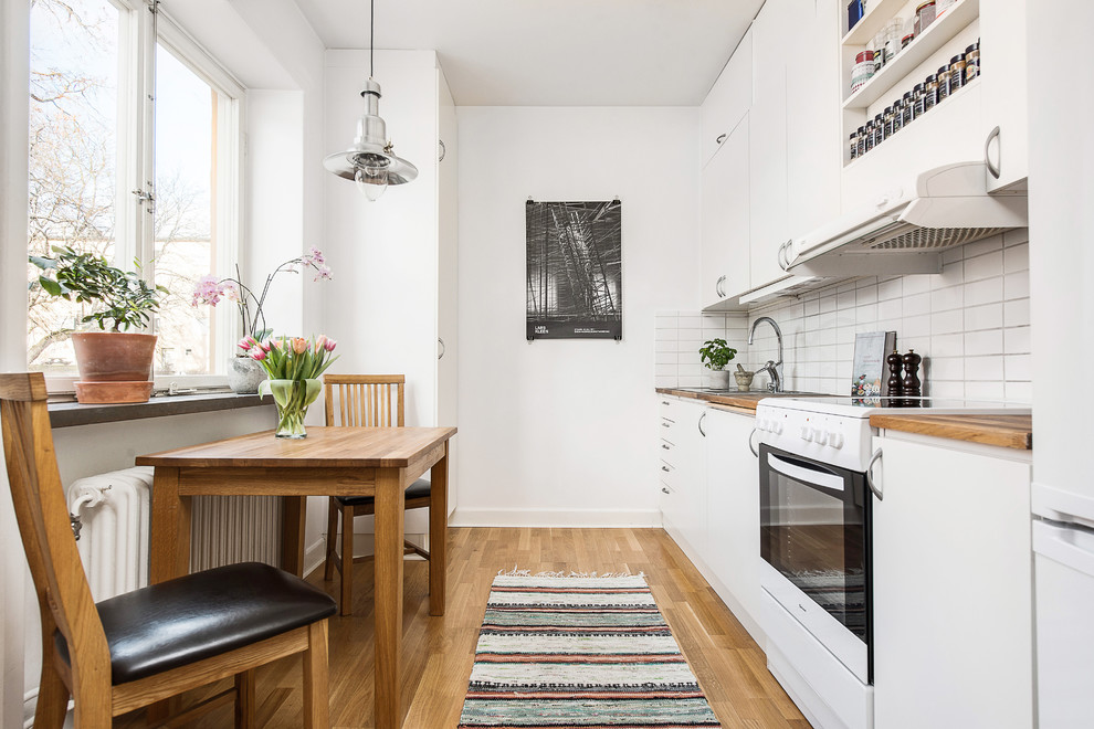 Example of a mid-sized danish single-wall medium tone wood floor eat-in kitchen design in Stockholm with flat-panel cabinets, white cabinets, white backsplash, white appliances, no island, wood countertops and ceramic backsplash
